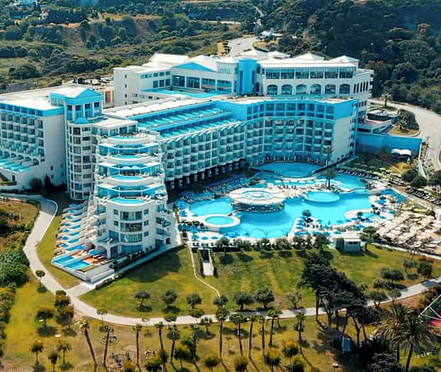 Suuri hotellirakennus, jossa on useita sinisiä parvekkeita ja uima-altaita, vehreyden ympäröimänä. (Tekoälyn luoma teksti)