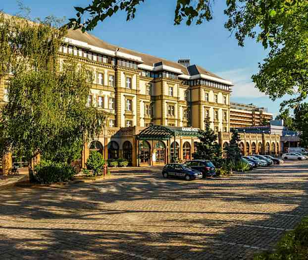 Kuva rakennuksesta, jonka julkisivussa lukee 'Grand Hotel', ympärillä puita ja pysäköityjä autoja. (Tekoälyn luoma teksti)