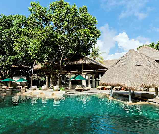 Ulkouima-allas, jossa on kirkasta vettä, ympäröitynä puilla ja olkikattoisilla rakenteilla. Aurinkovarjoja ja aurinkotuoleja on sijoitettu viereiselle terassille. (Tekoälyn luoma teksti)