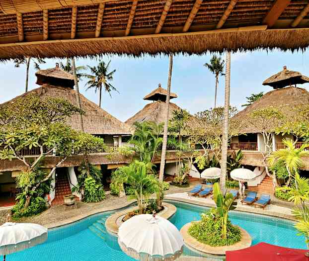 Lomahotelli, jossa on uima-allas, jota ympäröi palmuja ja olkikattoisia rakennuksia. (Tekoälyn luoma teksti)