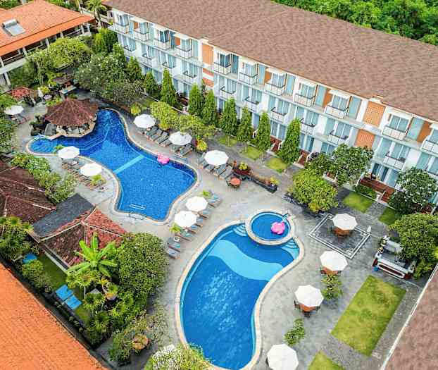 Lintuperspektiivistä otettu kuva hotellialueesta, jossa on kaksi uima-allasta, aurinkotuoleja ja varjoja, ympärillä kasvillisuutta ja rakennuksia. (Tekoälyn luoma teksti)