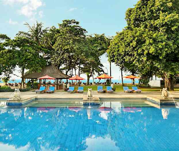 Ulkouima-allas, jota ympäröivät aurinkotuolit, aurinkovarjot ja puut, taustalla meri. (Tekoälyn luoma teksti)