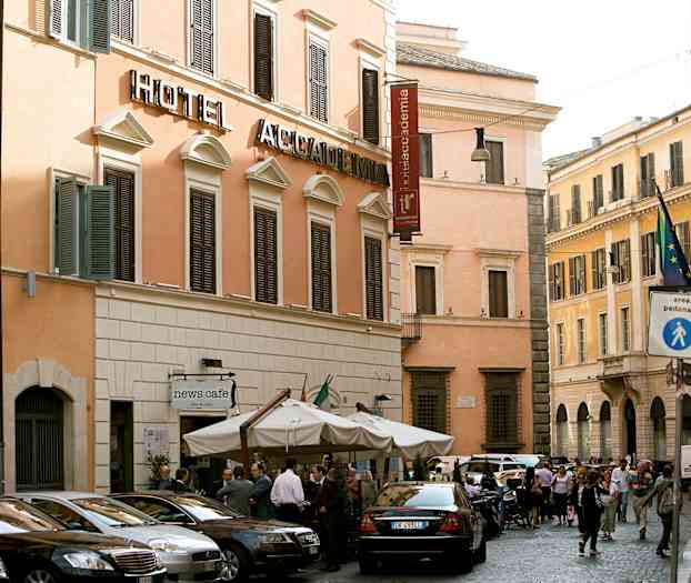 Katunäkymä, jossa hotellirakennus beigeillä julkisivuilla ja tummilla ikkunaluukuilla. Rakennuksen edessä on useita pysäköityjä autoja ja kahvila aurinkovarjoineen. (Tekoälyn luoma teksti)