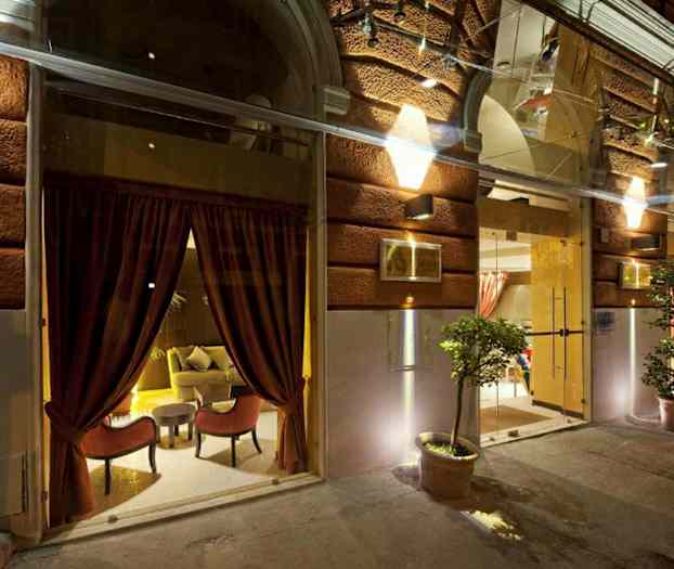 Kuva rakennuksen sisäänkäynnistä, jossa on valaistu julkisivu, suuret lasi-ikkunat punaisilla verhoilla ja ruukkukasveilla. (Tekoälyn luoma teksti)