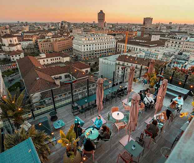 Kattoterassi, jolla on pöytiä ja istumapaikkoja, ympärillä panoraamanäkymä kaupunkiin auringonlaskun aikaan. (Tekoälyn luoma teksti)