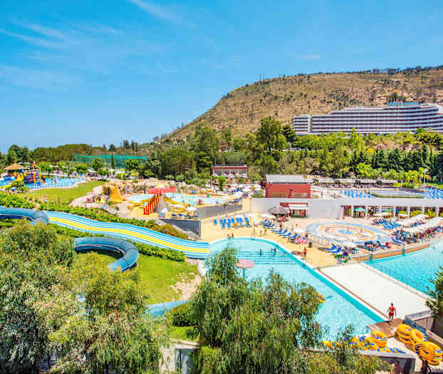 Kuvassa näkyy suuri ulkoilman vesipuisto, jossa on altaita, vesiliukumäkiä ja ympäröiviä aurinkotuoleja. Taustalla näkyy suuri rakennus ja kalteva maasto. (Tekoälyn luoma teksti)