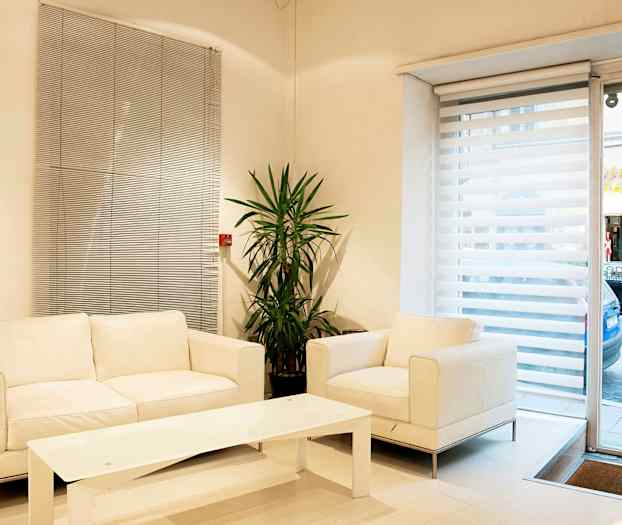 Vaalea olohuone, jossa valkoisia huonekaluja, kasvi ja suuret ikkunat sälekaihtimilla. (Tekoälyn luoma teksti)