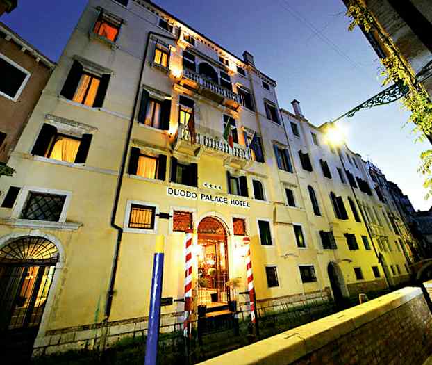 Kolmikerroksinen asuinrakennus, jossa kyltti Duodo Palace Hotel, kanavan ja kävelytien ympäröimänä. (Tekoälyn luoma teksti)