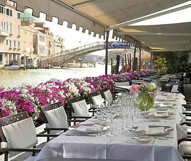 Katettu ruokapöytä valkoisilla liinoilla, jotka on asetettu aurinkokatoksen alle kanavan varrelle, joka on täynnä veneitä ja reunustettu värikkäillä kukilla. (Tekoälyn luoma teksti)