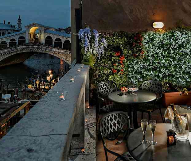 Kuvassa on terassi, jossa on pöytiä ja tuoleja, näkymä Venedigin Rialton sillalle sekä kukkia ja kynttilöitä koristelemassa aluetta. (Tekoälyn luoma teksti)