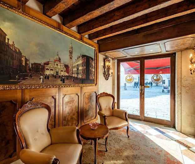 Sisätilakohtaus, jossa kaksi tuolia ja pöytä maalauksella koristellun seinän edessä, näkymä ulkotiloihin lasiovien kautta. (Tekoälyn luoma teksti)