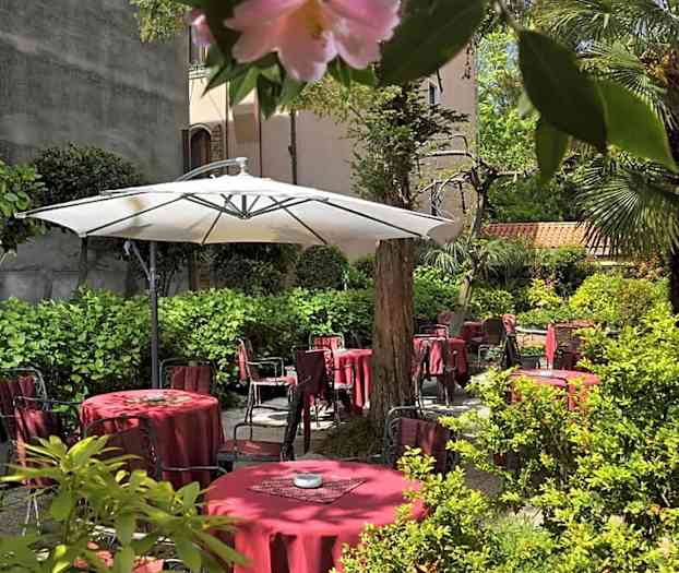 Vehreä sisäpiha, jossa punaisia pöytiä ja tuoleja sekä suuri valkoinen varjo. (Tekoälyn luoma teksti)