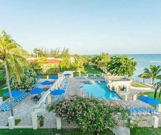 Kuva hotellialueesta meren äärellä, jossa on uima-allas, aurinkotuoleja ja aurinkovarjoja vehreyden ja palmujen ympäröimänä. (Tekoälyn luoma teksti)