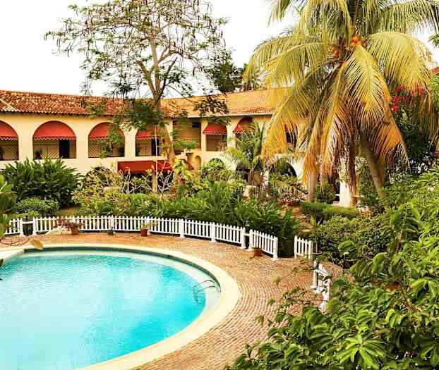 Pyöreä uima-allas, jota ympäröi kivipäällyste ja valkoiset aidat. Taustalla on rakennus, jossa on holvaikkunat ja punaiset markiisit sekä ympärillä vihreää kasvillisuutta. (Tekoälyn luoma teksti)