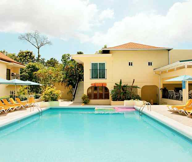 Uima-allas, jossa ympärillä aurinkotuoleja ja aurinkovarjoja. Altaan takana on rakennus, jossa on vaaleat seinät ja terrakotan värinen kattotiili. (Tekoälyn luoma teksti)