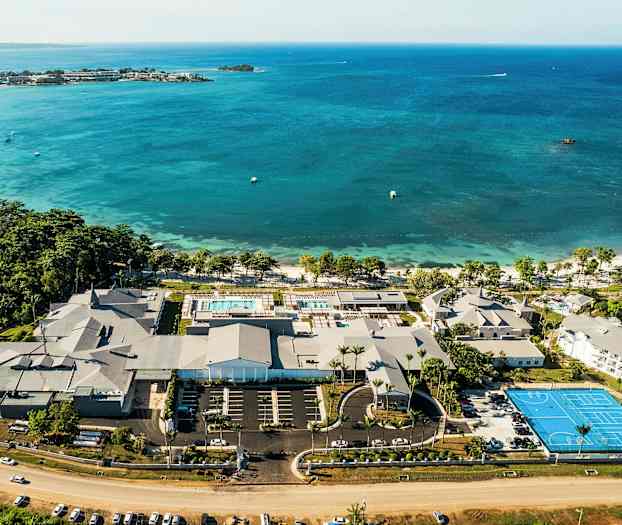 Ilmakuva rantalomakohteesta, jossa on uima-altaita, rakennuksia ja tenniskenttä meren rannalla. (Tekoälyn luoma teksti)