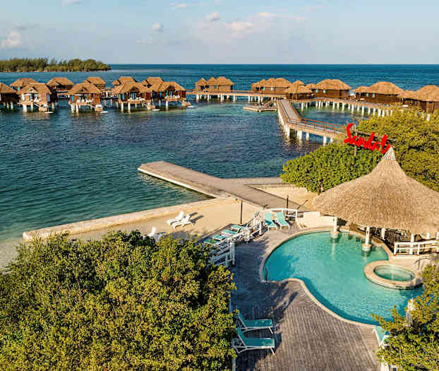 Merelle rakennettuja pylväiden päällä olevia bungaloweja, joissa etualalla uima-allas ja aurinkotuoleja. (Tekoälyn luoma teksti)