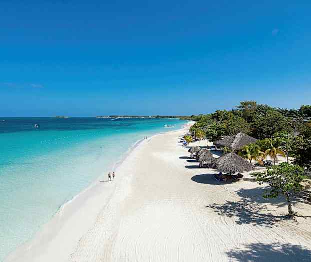 Valkoinen hiekkaranta, jossa on aurinkovarjoja ja kirkasta sinistä vettä, ympärillä vehreyttä. (Tekoälyn luoma teksti)