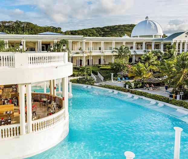 Suuri rakennus valkoisine pylväineen ja kupoleineen uima-altaan äärellä, ympärillä palmuja ja aurinkotuoleja. (Tekoälyn luoma teksti)