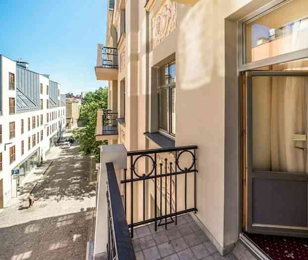 Kuvassa näkyy parveke vaaleassa rakennuksessa, josta on näkymä mukulakivikadulle ja moderniin rakennukseen toisella puolella katua. (Tekoälyn luoma teksti)