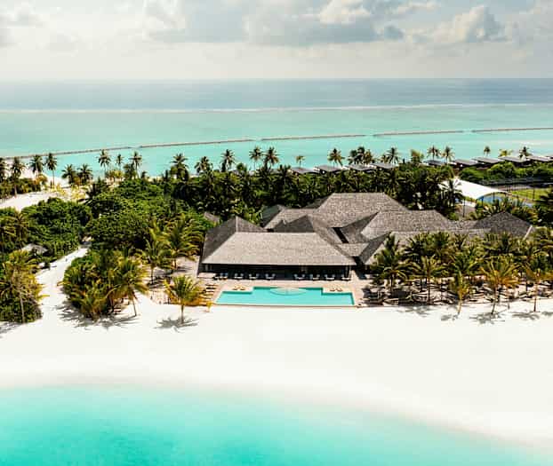 Ilmakuva hiekkarannasta, jossa on uima-allas, palmupuita ja rakennuksia meren äärellä. (Tekoälyn luoma teksti)