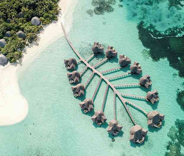 Ilmakuva bungaloweista pylväiden päällä kirkkaan sinisen veden yllä, lähellä hiekkarantaa ja vehreää kasvillisuutta. (Tekoälyn luoma teksti)