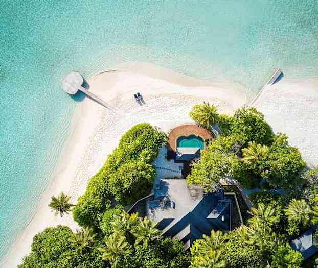 Ilmakuva hiekkarannasta kirkkaan trooppisen meren äärellä, rehevällä kasvillisuudella ja pienellä rakennuksella. (Tekoälyn luoma teksti)