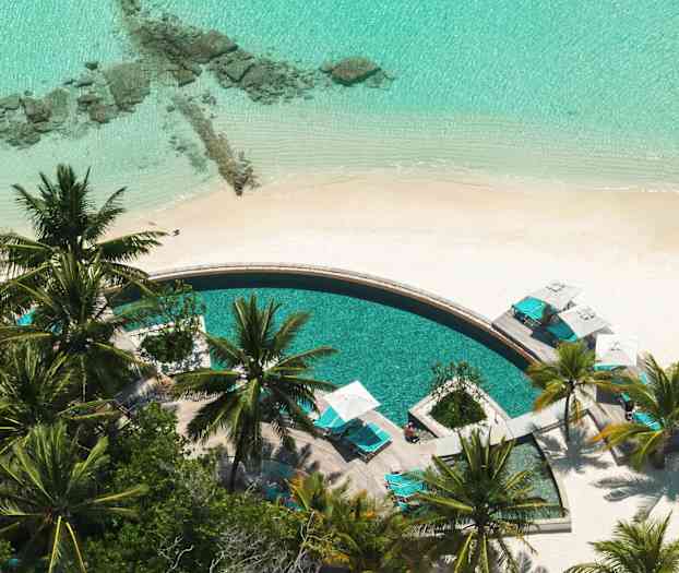 Pyöreä uima-allas, jota ympäröivät aurinkotuolit ja palmut hiekkarannalla. (Tekoälyn luoma teksti)