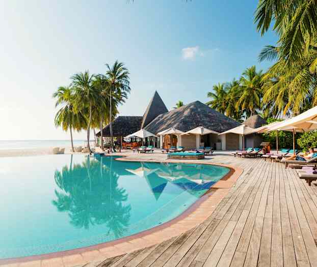 Infiniteettiuima-allas, jonka reunalla on puita ja aurinkovarjoja, taustalla olkikattoisia rakennuksia. (Tekoälyn luoma teksti)