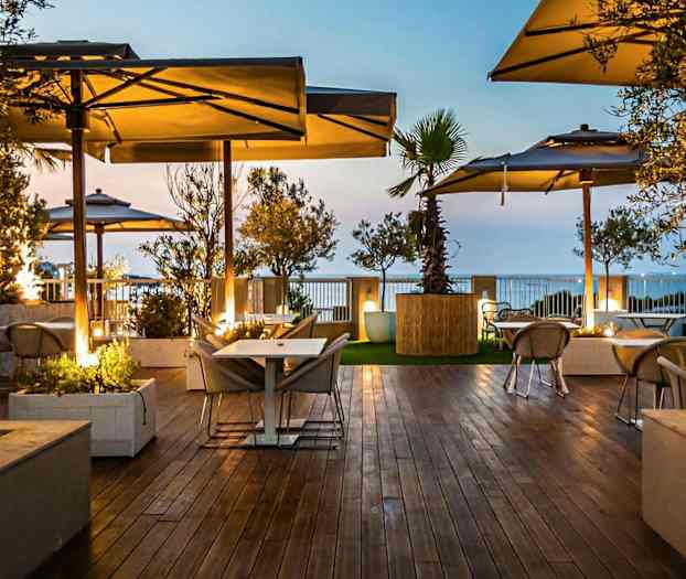 Kuva ulkoterassista, jossa puiset lattiat, pöytiä ja tuoleja aurinkovarjojen alla, vehreyden ympäröimänä iltavalossa. (Tekoälyn luoma teksti)