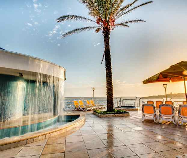 Uima-allas, jossa on vesiputous palmun vieressä, aurinkotuoleja ja näkymä merelle aurinkoisena päivänä. (Tekoälyn luoma teksti)