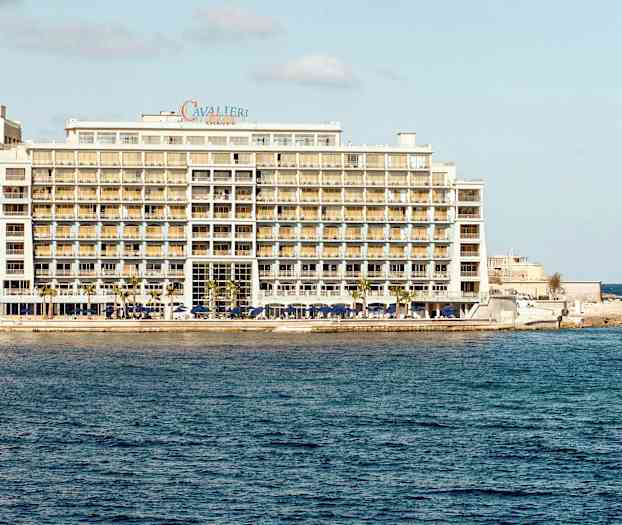 Suuri hotelli meren rannalla, jossa on useita kerroksia ja parvekkeita, sekä pieni vene vedessä lähellä hotellia. (Tekoälyn luoma teksti)