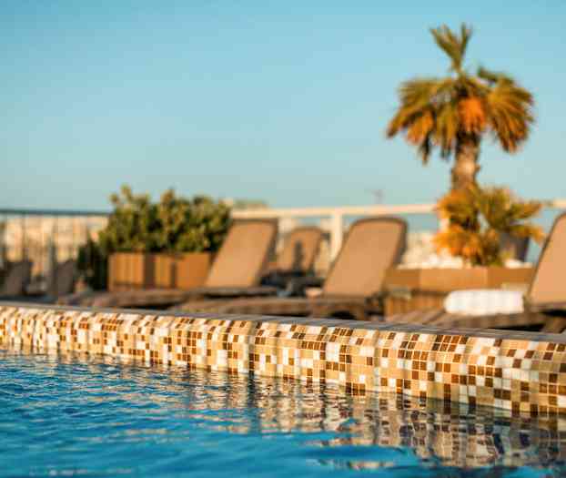 Lähikuva uima-altaan reunasta kirkkaalla vedellä, taustalla aurinkotuoleja ja kasveja. (Tekoälyn luoma teksti)