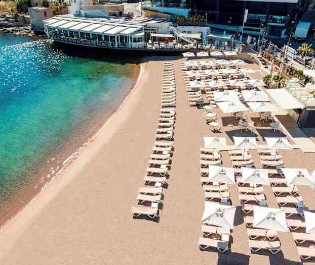 Ranta, jossa aurinkotuoleja ja aurinkovarjoja sekä lähellä oleva rakennus. (Tekoälyn luoma teksti)