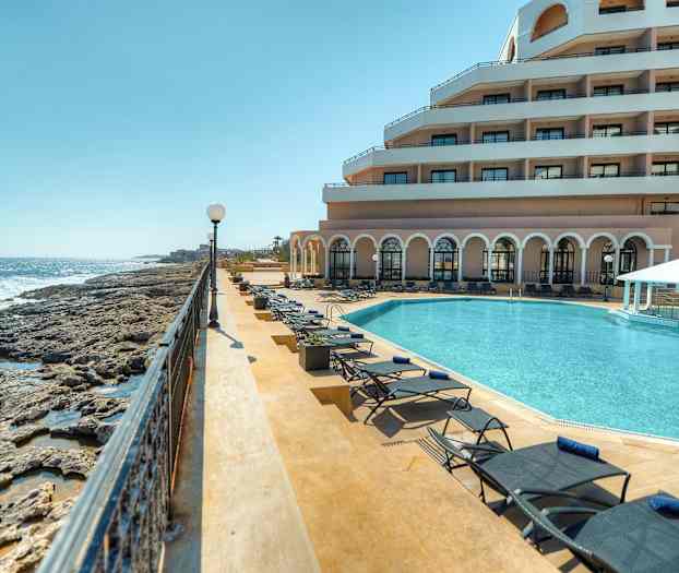 Kuva ylellisestä lomakeskuksesta meren rannalla, jossa on uima-allas, aurinkotuoleja ja parvekkeellinen rakennus. (Tekoälyn luoma teksti)