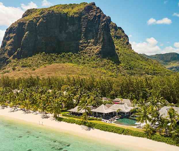 Taustalla näkyy suuri vuori, ja etualalla on ranta, jossa on turkoosia vettä. Rannalla on palmuja ja olkikattoisia rakennuksia. (Tekoälyn luoma teksti)