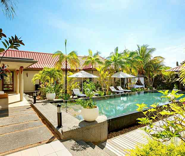 Avoin ympäristö, jossa uima-allas, ympäröitynä aurinkotuoleilla ja varjoilla, vehreyden ja rakennusten ympäröimänä. (Tekoälyn luoma teksti)