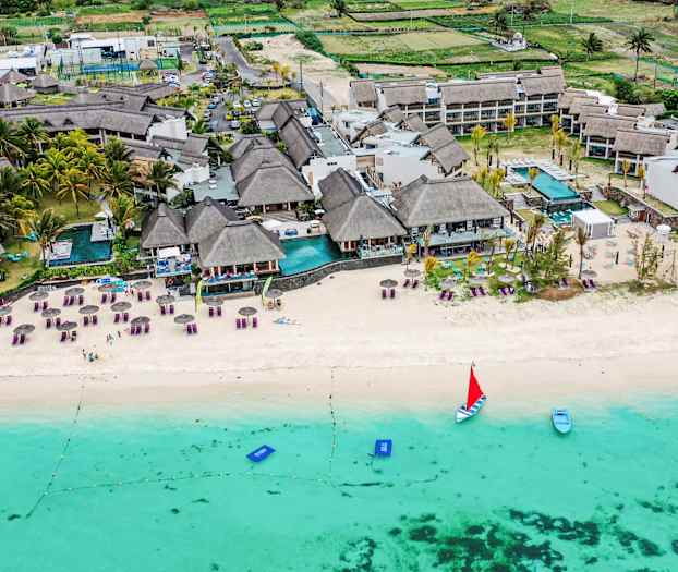 Ilmakuva rannasta, jossa on aurinkotuoleja, varjoja ja lomakeskus vesirajassa. (Tekoälyn luoma teksti)