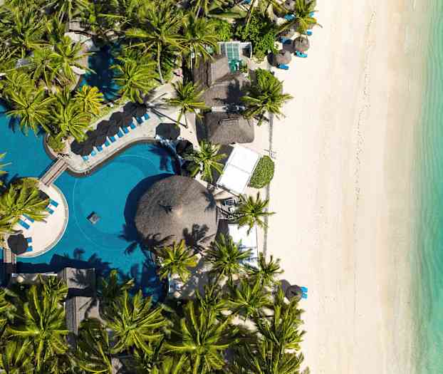 Kuva esittää ilmakuvaa rannasta, jossa on valkoista hiekkaa, turkoosia vettä ja palmun ympäröimä uima-allas. (Tekoälyn luoma teksti)