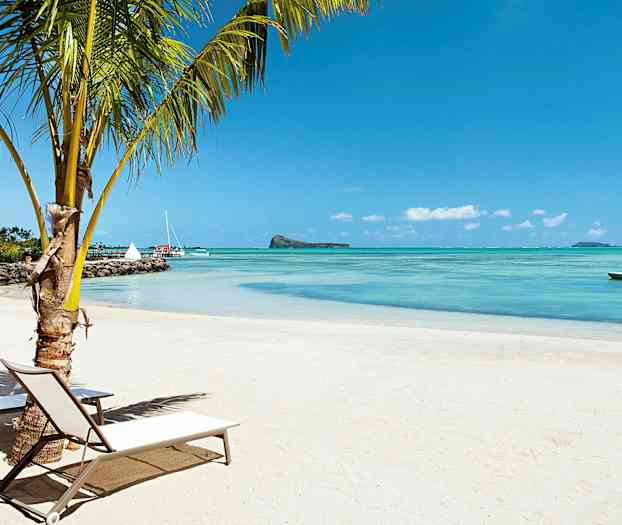 Hiekkaranta, jossa on palmu ja aurinkotuoleja veden äärellä. Taustalla näkyy purjeveneitä ja pieni saari. (Tekoälyn luoma teksti)