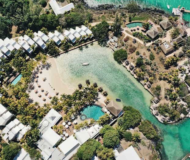 Ilmakuva hotellialueesta, jossa on uima-allas, hiekkaranta ja vehreyttä veden äärellä. (Tekoälyn luoma teksti)