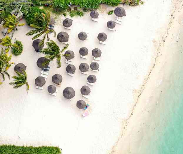 Ilmakuva hiekkarannasta, jossa aurinkotuoleja ja varjoja lähellä vettä ja kasvillisuutta. (Tekoälyn luoma teksti)