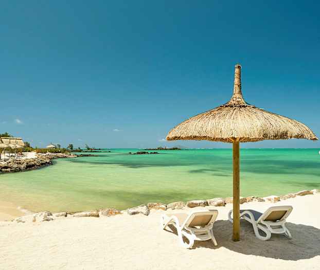 Hiekkaranta, jossa on kaksi aurinkotuolia olkikattoisen päivänvarjon alla, taustalla turkoosia vettä. (Tekoälyn luoma teksti)