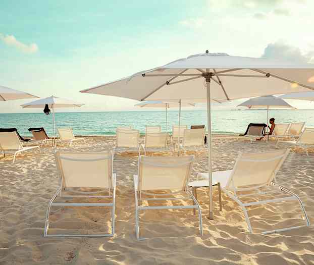 Hiekkaranta, jossa on aurinkotuoleja ja aurinkovarjoja. Taustalla näkyy meri kirkkaan taivaan alla. (Tekoälyn luoma teksti)