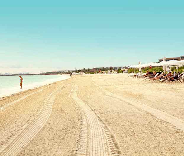 Hiekkaranta, jossa on aurinkovarjoja ja aurinkotuoleja, yksi henkilö seisoo veden äärellä. (Tekoälyn luoma teksti)