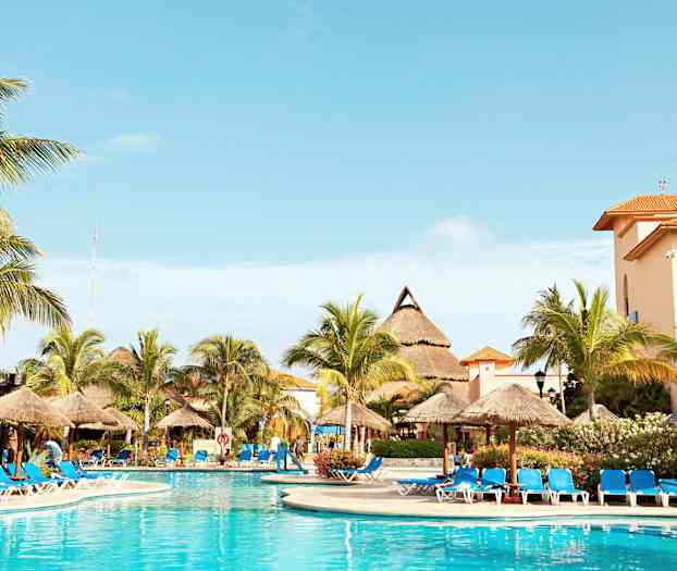 Kuvassa näkyy ulkouima-allas, jota ympäröivät sinisillä tyynyillä varustetut aurinkotuolit ja aurinkovarjot. Taustalla on palmuja ja rakennuksia oranssinsävyisine kattoineen. (Tekoälyn luoma teksti)