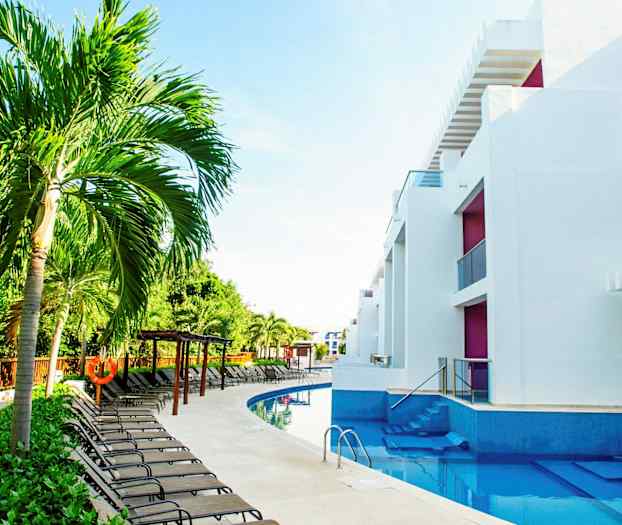 Kuva uima-altaasta, jonka ympärillä on aurinkotuoleja, palmuja ja moderneja valkoisia rakennuksia. (Tekoälyn luoma teksti)