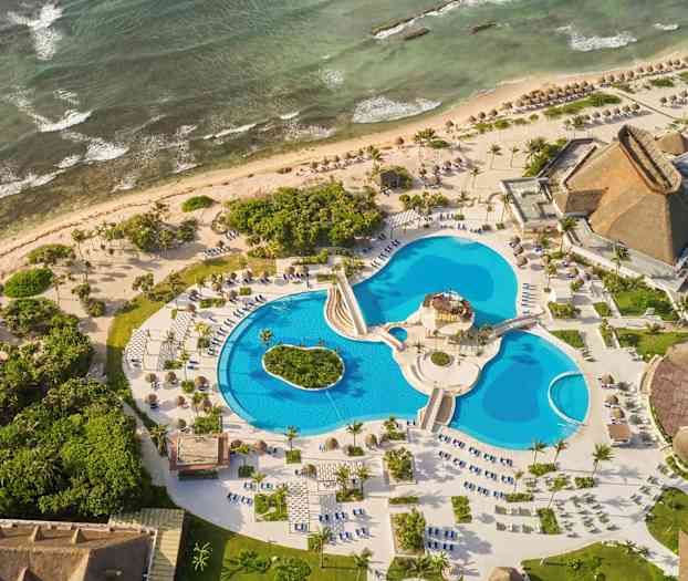 Ilmakuva lomakeskuksesta meren rannalla, jossa on useita uima-altaita, ympäröivää vehreyttä ja rakennuksia olkikatolla. (Tekoälyn luoma teksti)