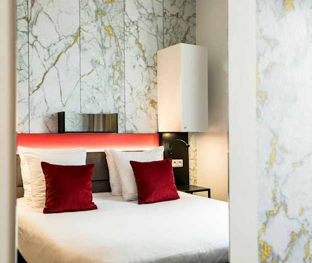 Moderni hotellihuone, jossa on parisänky, valkoisia ja punaisia tyynyjä sekä taustaseinässä marmorikuvio. (Tekoälyn luoma teksti)