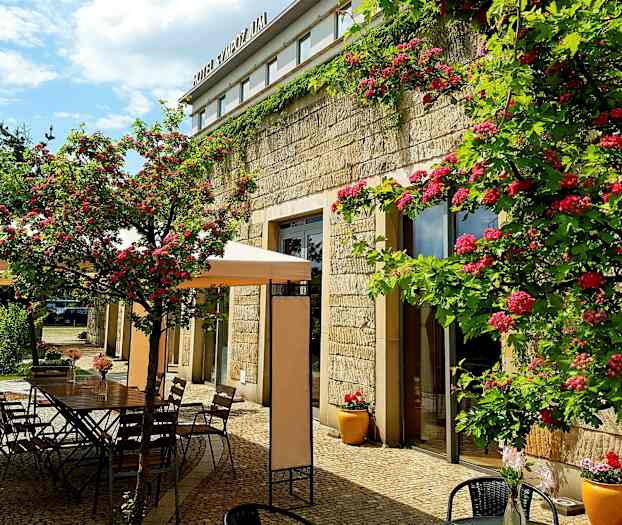 Ulkoiluterassi kukkien ja pöytien kanssa kivirakennuksen vieressä. (Tekoälyn luoma teksti)